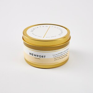 May include: A gold-colored candle tin with a white and beige label. The label reads "NEWPORT candle" and "SCRIPTED FRAGRANCE". The tin has a lid and contains text describing the scent. A natural soy candle.