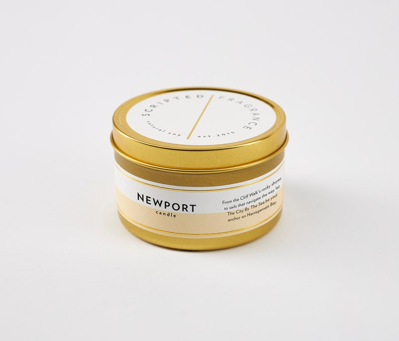 May include: A gold-colored candle tin with a white and beige label. The label reads "NEWPORT candle" and "SCRIPTED FRAGRANCE". The tin has a lid and contains text describing the scent. A natural soy candle.