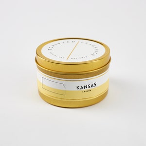 K&ouml;nnte beinhalten: Goldene Dose mit einer Kerze und einem wei&szlig;en Etikett mit der Aufschrift "Scripted Fragrance est. 2018" und "KANSAS candle". Das Etikett zeigt au&szlig;erdem eine Grafik des Bundesstaates Kansas.