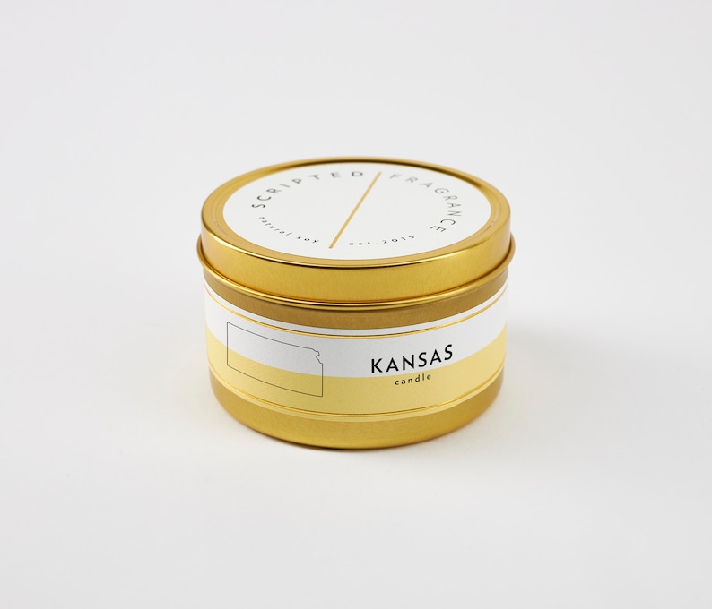 K&ouml;nnte beinhalten: Goldene Dose mit einer Kerze und einem wei&szlig;en Etikett mit der Aufschrift "Scripted Fragrance est. 2018" und "KANSAS candle". Das Etikett zeigt au&szlig;erdem eine Grafik des Bundesstaates Kansas.