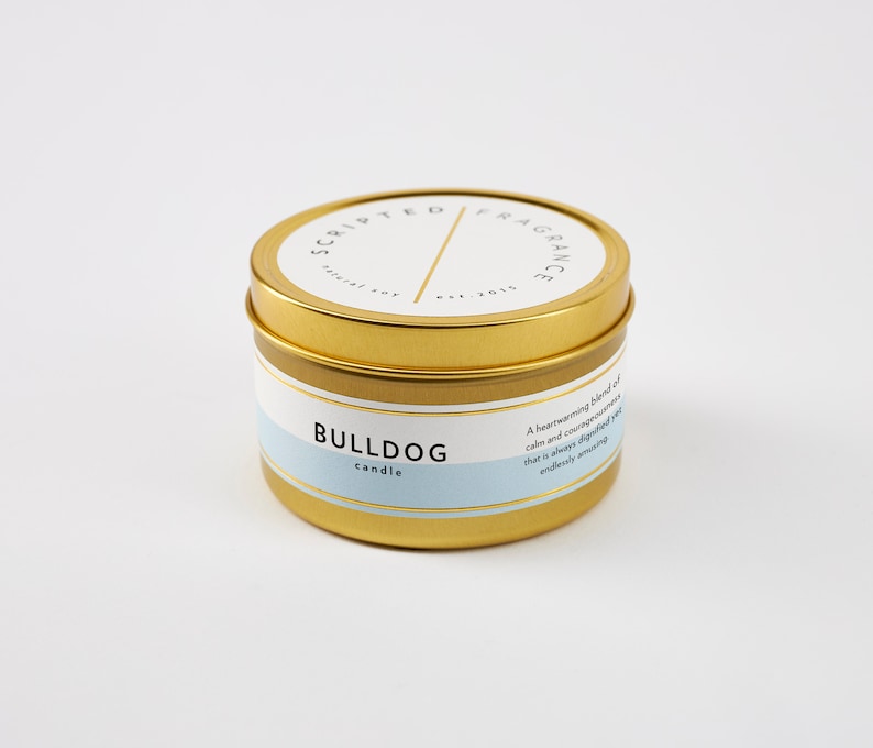Puede incluir: Vela en lata dorada con una etiqueta azul y blanca que dice "Bulldog candle". La etiqueta tambi&eacute;n incluye el texto "Una mezcla reconfortante de calma y valent&iacute;a que siempre es digna pero infinitamente encantadora."