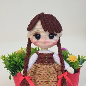 May include: Crocheted doll with brown hair in braids, wearing a brown and tan dress with a white blouse. The doll has a pink face and is standing in a red planter with greenery and flowers. The text "Amiville" is at the bottom.