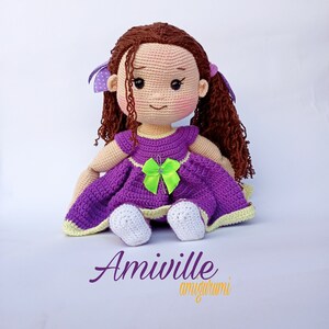 Puede incluir: Mu&ntilde;eca amigurumi Amiiville. Esta mu&ntilde;eca hecha a mano tiene cabello casta&ntilde;o rizado, una cara sonriente con mejillas rosadas y viste un vestido morado con un lazo verde. La mu&ntilde;eca tiene zapatos blancos y lazos morados en el pelo. La mu&ntilde;eca est&aacute; sentada.