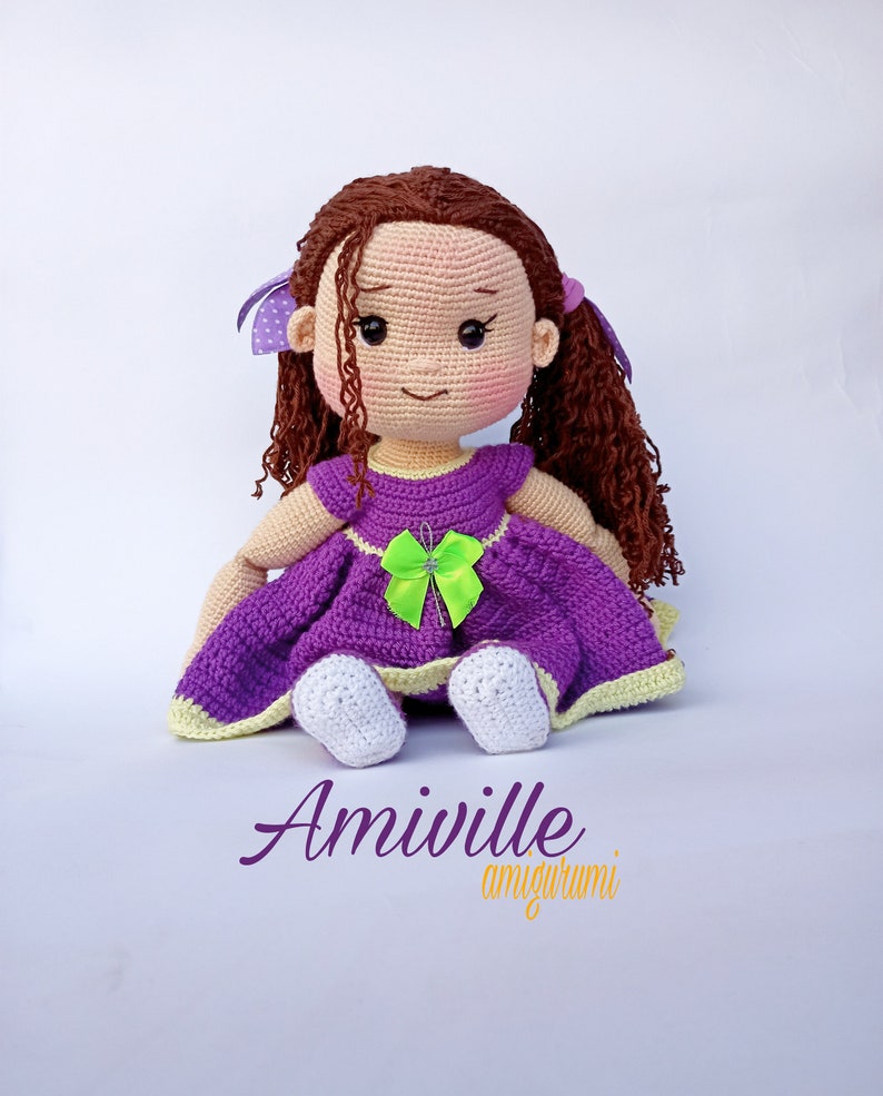 Puede incluir: Mu&ntilde;eca amigurumi Amiiville. Esta mu&ntilde;eca hecha a mano tiene cabello casta&ntilde;o rizado, una cara sonriente con mejillas rosadas y viste un vestido morado con un lazo verde. La mu&ntilde;eca tiene zapatos blancos y lazos morados en el pelo. La mu&ntilde;eca est&aacute; sentada.
