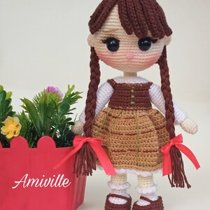 May include: Crocheted doll with brown hair in braids, wearing a brown and tan dress with red bows. The doll has a white shirt and brown shoes. A red planter with flowers is next to the doll.