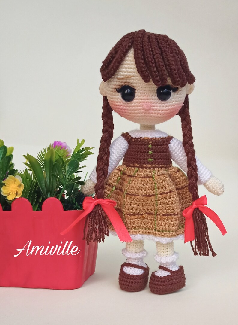 May include: Crocheted doll with brown hair in braids, wearing a brown and tan dress with red bows. The doll has a white shirt and brown shoes. A red planter with flowers is next to the doll.