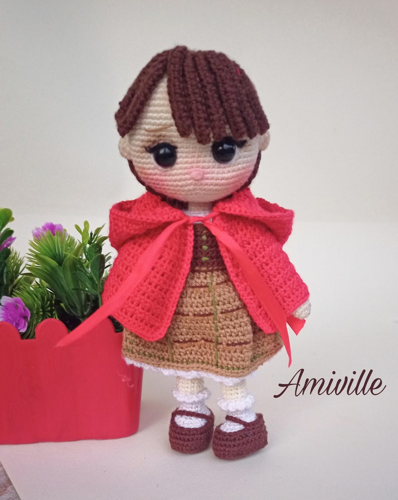 May include: Amiiville crochet doll of a girl wearing a red hooded cape and brown skirt. The doll has brown hair, large black eyes, and is wearing burgundy shoes. A red flower pot is in the background.