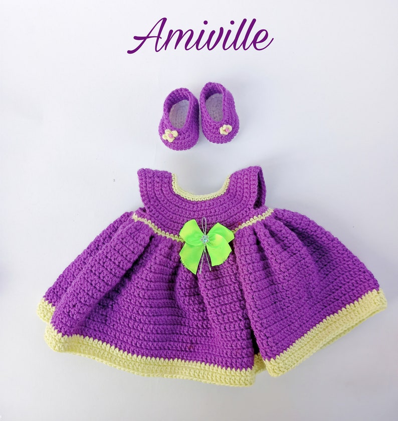 Puede incluir: Amiwille. Conjunto de vestido y zapatos de beb&eacute; de ganchillo, morado con ribete amarillo y lazo verde brillante. El conjunto incluye un vestido y zapatos a juego. El texto "Amiwille" est&aacute; en la esquina superior izquierda.