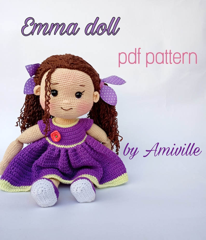 Puede incluir: Mu&ntilde;eca de ganchillo hecha a mano llamada "Emma", con un vestido morado con ribetes amarillos y una flor roja. La mu&ntilde;eca tiene cabello rizado casta&ntilde;o, lazos morados con lunares y zapatos blancos. El texto "pdf pattern by Amiville" es visible.