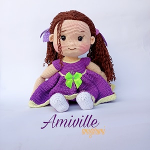 Puede incluir: Mu&ntilde;eca de ganchillo hecha a mano con cabello casta&ntilde;o rizado, vestida con un vestido morado con un lazo verde. La mu&ntilde;eca tiene mejillas rosadas y est&aacute; sentada. El texto "Amiville amigurumi" est&aacute; escrito debajo.