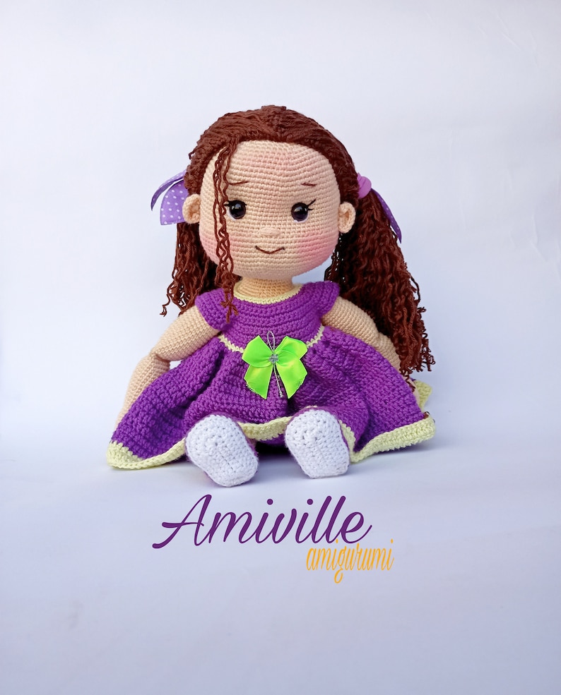 Puede incluir: Mu&ntilde;eca de ganchillo hecha a mano con cabello casta&ntilde;o rizado, vestida con un vestido morado con un lazo verde. La mu&ntilde;eca tiene mejillas rosadas y est&aacute; sentada. El texto "Amiville amigurumi" est&aacute; escrito debajo.