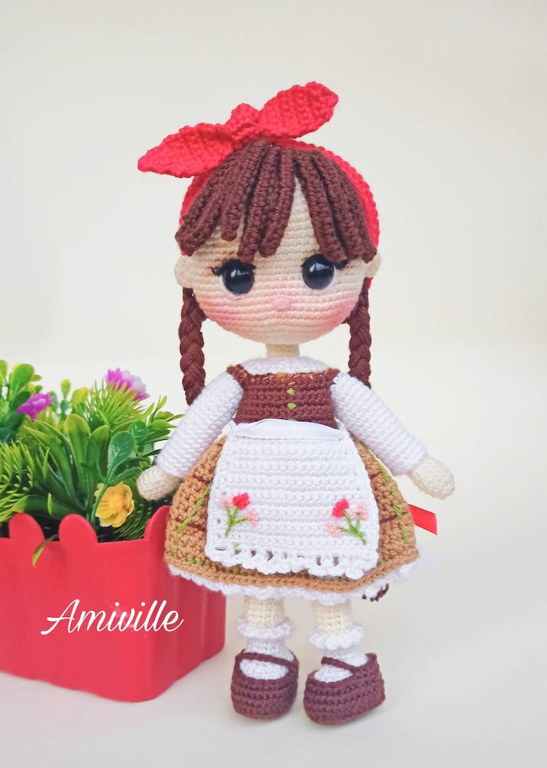 May include: Crocheted doll with brown hair in braids, wearing a red bow, white blouse, brown vest, and a floral-embroidered apron. The doll has dark eyes and is wearing brown shoes. The doll is next to a red planter with flowers. The word "Amiville" is on the planter.