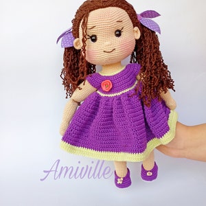 Puede incluir: Una mu&ntilde;eca de ganchillo hecha a mano con cabello rizado casta&ntilde;o, vestida con un vestido morado con un ribete verde claro y una flor rosa. La mu&ntilde;eca tiene zapatos y lazos morados. La palabra "Amiville" est&aacute; escrita en una fuente lila claro.