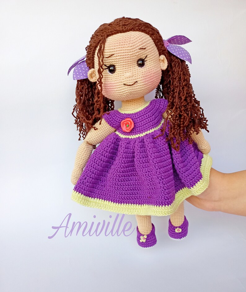 Puede incluir: Una mu&ntilde;eca de ganchillo hecha a mano con cabello rizado casta&ntilde;o, vestida con un vestido morado con un ribete verde claro y una flor rosa. La mu&ntilde;eca tiene zapatos y lazos morados. La palabra "Amiville" est&aacute; escrita en una fuente lila claro.