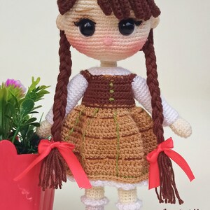 May include: A handmade crochet doll with brown hair in braids, wearing a brown and tan dress with a white blouse. The doll has black eyes and is wearing brown shoes. A red pot with flowers is on the left. The text "Amiville" is on the right.