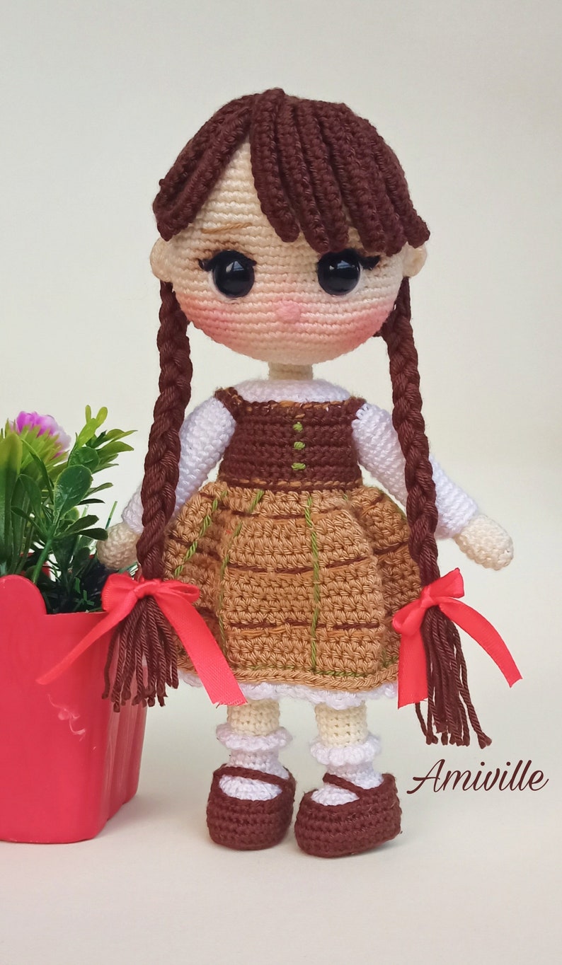 May include: A handmade crochet doll with brown hair in braids, wearing a brown and tan dress with a white blouse. The doll has black eyes and is wearing brown shoes. A red pot with flowers is on the left. The text "Amiville" is on the right.
