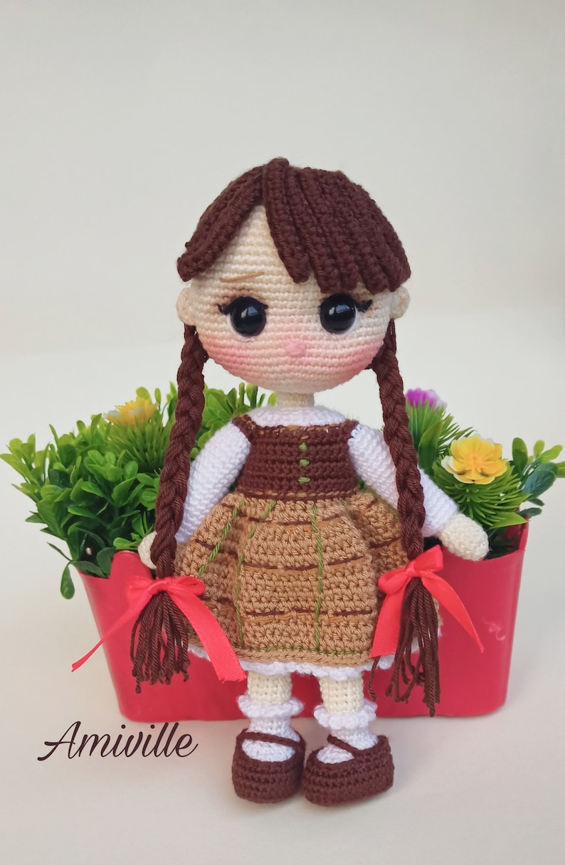 May include: Crocheted doll with brown hair in braids, wearing a brown and tan dress with a white blouse. The doll has a pink face and is standing in a red planter with greenery and flowers. The text "Amiville" is at the bottom.