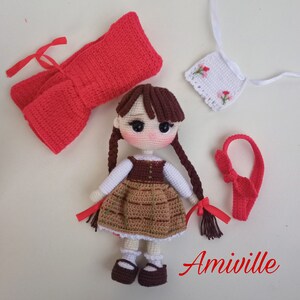 May include: Crocheted doll with brown hair in pigtails, wearing a brown and tan dress, white undershirt, and brown shoes. Accessories include a red bow, a red headband, and a white embroidered apron. The word "Amiville" is written in red.