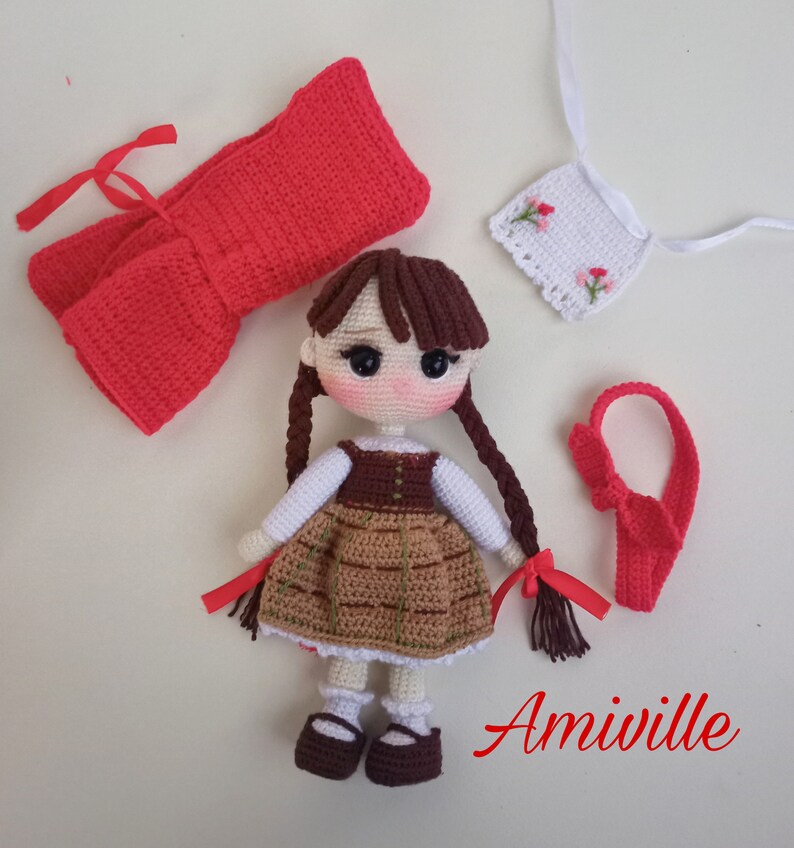 May include: Crocheted doll with brown hair in pigtails, wearing a brown and tan dress, white undershirt, and brown shoes. Accessories include a red bow, a red headband, and a white embroidered apron. The word "Amiville" is written in red.