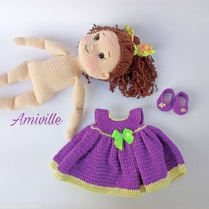 Puede incluir: Mu&ntilde;eca de ganchillo con cabello casta&ntilde;o rizado, tez clara y cuerpo de ganchillo a juego. La mu&ntilde;eca va acompa&ntilde;ada de un vestido morado con un lazo verde y zapatos morados. La palabra "Amiville" est&aacute; escrita en p&uacute;rpura.
