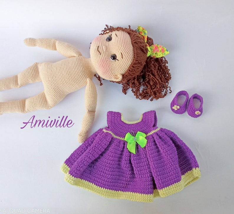 Puede incluir: Mu&ntilde;eca de ganchillo con cabello casta&ntilde;o rizado, tez clara y cuerpo de ganchillo a juego. La mu&ntilde;eca va acompa&ntilde;ada de un vestido morado con un lazo verde y zapatos morados. La palabra "Amiville" est&aacute; escrita en p&uacute;rpura.