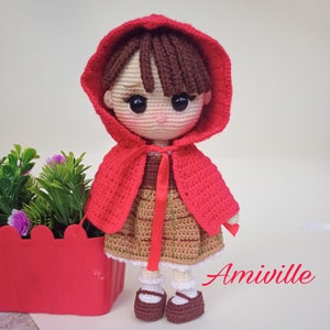 May include: Amiwille crochet doll wearing a red hooded cape and a brown dress with a tan skirt. The doll has brown hair and is wearing brown shoes. The doll is standing next to a red planter with green plants and purple flowers.
