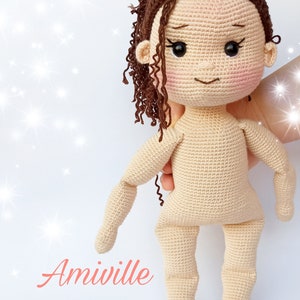 Puede incluir: Mu&ntilde;eca de ganchillo con cabello rizado casta&ntilde;o y un lazo floral. La mu&ntilde;eca tiene mejillas rosadas y no lleva ropa. La palabra "Amiville" est&aacute; escrita en cursiva en la parte inferior izquierda de la imagen.
