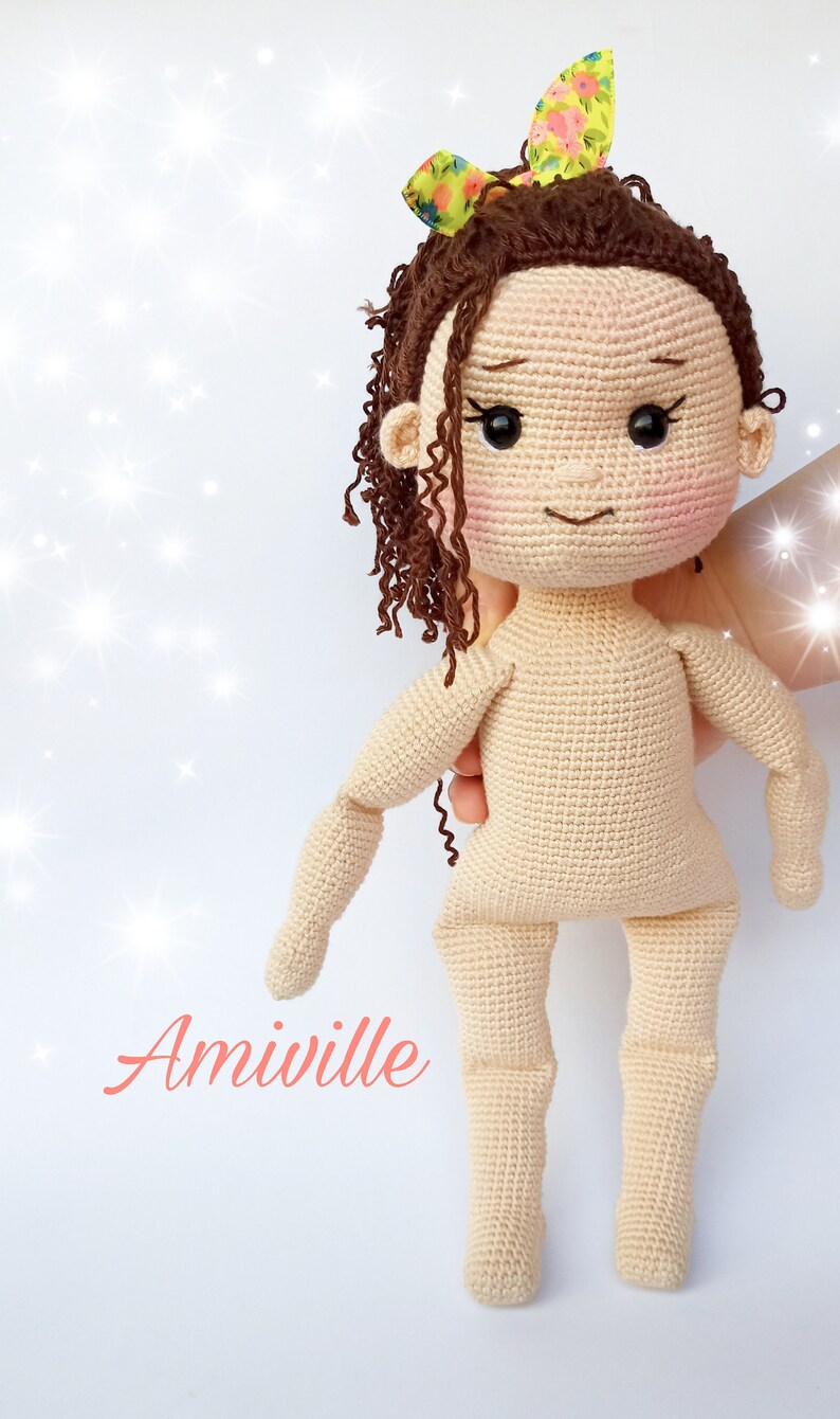 Puede incluir: Mu&ntilde;eca de ganchillo con cabello rizado casta&ntilde;o y un lazo floral. La mu&ntilde;eca tiene mejillas rosadas y no lleva ropa. La palabra "Amiville" est&aacute; escrita en cursiva en la parte inferior izquierda de la imagen.