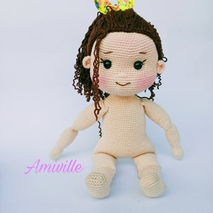 Puede incluir: Mu&ntilde;eca de ganchillo con cabello rizado casta&ntilde;o y un lazo floral amarillo. La mu&ntilde;eca tiene un tono de piel claro, mejillas rosadas y ojos negros. Los brazos y las piernas de la mu&ntilde;eca est&aacute;n extendidos, y el texto "Amiville" es visible.