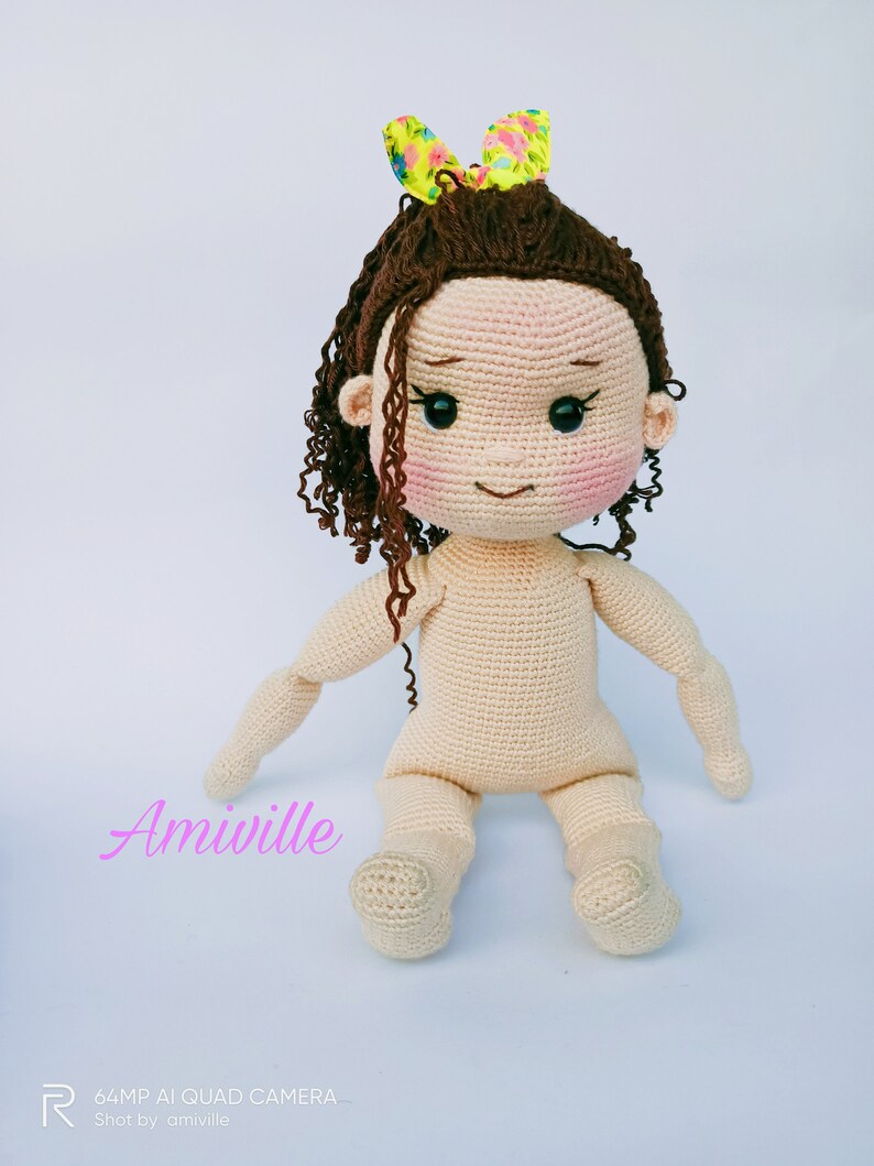 Puede incluir: Mu&ntilde;eca de ganchillo con cabello rizado casta&ntilde;o y un lazo floral amarillo. La mu&ntilde;eca tiene un tono de piel claro, mejillas rosadas y ojos negros. Los brazos y las piernas de la mu&ntilde;eca est&aacute;n extendidos, y el texto "Amiville" es visible.