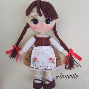 May include: A handmade crochet doll with brown braids tied with red ribbons. The doll wears a white blouse, a brown vest, and a skirt with floral embroidery. The doll has rosy cheeks and is wearing brown shoes. The text "Amiville" is visible.