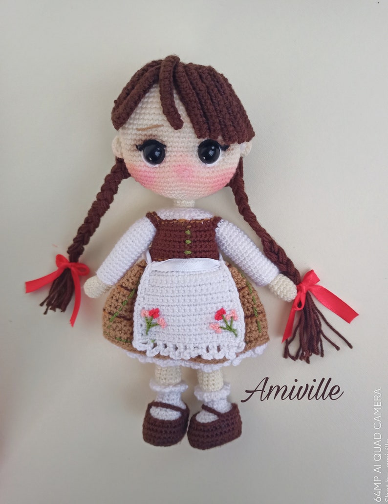 May include: A handmade crochet doll with brown braids tied with red ribbons. The doll wears a white blouse, a brown vest, and a skirt with floral embroidery. The doll has rosy cheeks and is wearing brown shoes. The text "Amiville" is visible.