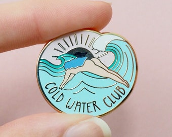 May include: A circular enamel pin with a gold edge. The pin depicts a swimmer diving into a wave, with the words "COLD WATER CLUB" arranged in a circle. The design incorporates blue, white, and black colours.