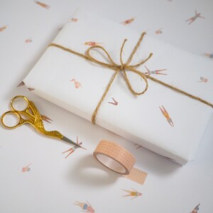 K&ouml;nnte beinhalten: Ein wei&szlig;es Geschenkpapier mit einer braunen Kordelschleife und einem sich wiederholenden Muster von Schwimmern. Eine goldene Schere und eine Rolle braunes, gemustertes Washi-Tape sind ebenfalls im Bild zu sehen.