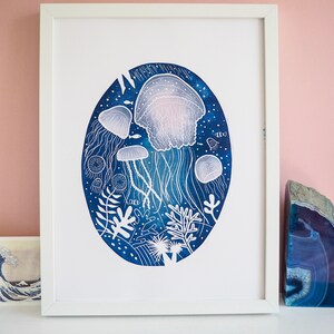 May include: A framed art print featuring a blue and white illustration of jellyfish and marine life. The artwork is set against a dark blue oval background with white accents, and is framed in a white rectangular frame.