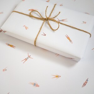 K&ouml;nnte beinhalten: Wei&szlig;e Geschenkpapier mit einem sich wiederholenden Muster aus braunen Strichm&auml;nnchen in Badeanz&uuml;gen. Das Geschenkpapier ist mit einer braunen Kordelschleife gebunden.