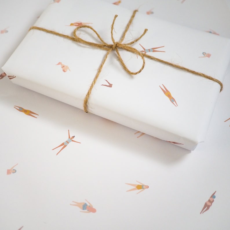 K&ouml;nnte beinhalten: Wei&szlig;e Geschenkpapier mit einem sich wiederholenden Muster aus braunen Strichm&auml;nnchen in Badeanz&uuml;gen. Das Geschenkpapier ist mit einer braunen Kordelschleife gebunden.
