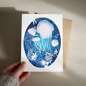 May include: A white greeting card featuring an oval illustration of jellyfish in a deep blue ocean. The jellyfish are white and light pink, with long, flowing tentacles. White coral and other sea life are also depicted.