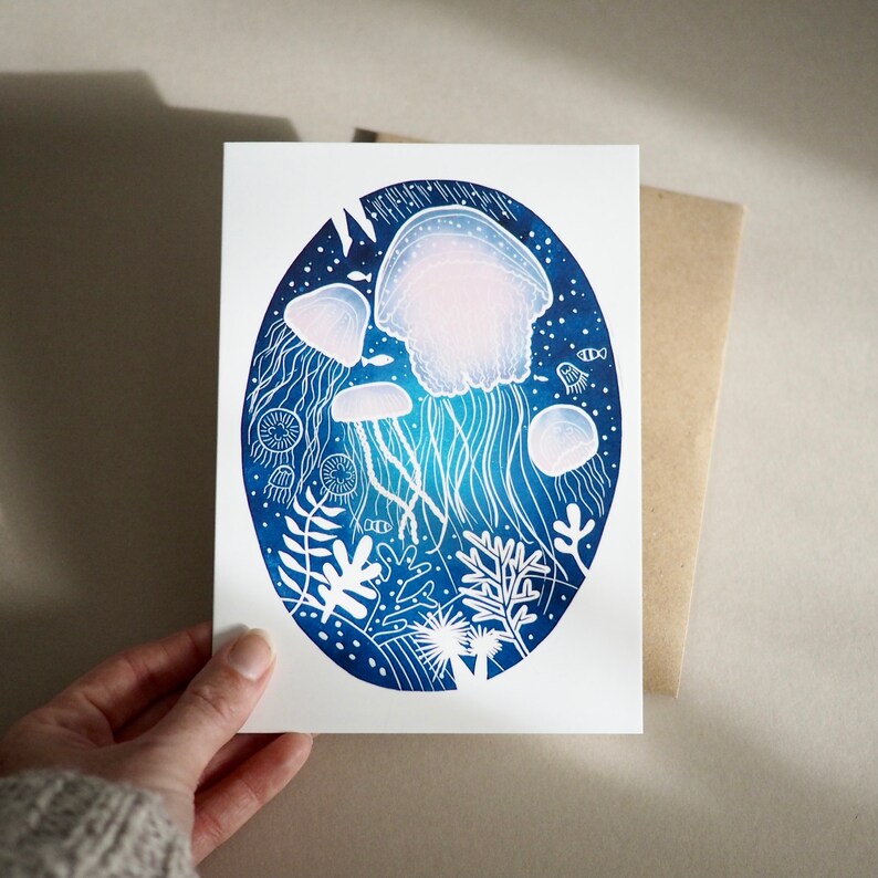 May include: A white greeting card featuring an oval illustration of jellyfish in a deep blue ocean. The jellyfish are white and light pink, with long, flowing tentacles. White coral and other sea life are also depicted.