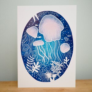 May include: An art print featuring a watercolor illustration of jellyfish in a deep blue ocean scene. The jellyfish are depicted in white and light pink, with long, flowing tentacles. The print includes coral and fish.