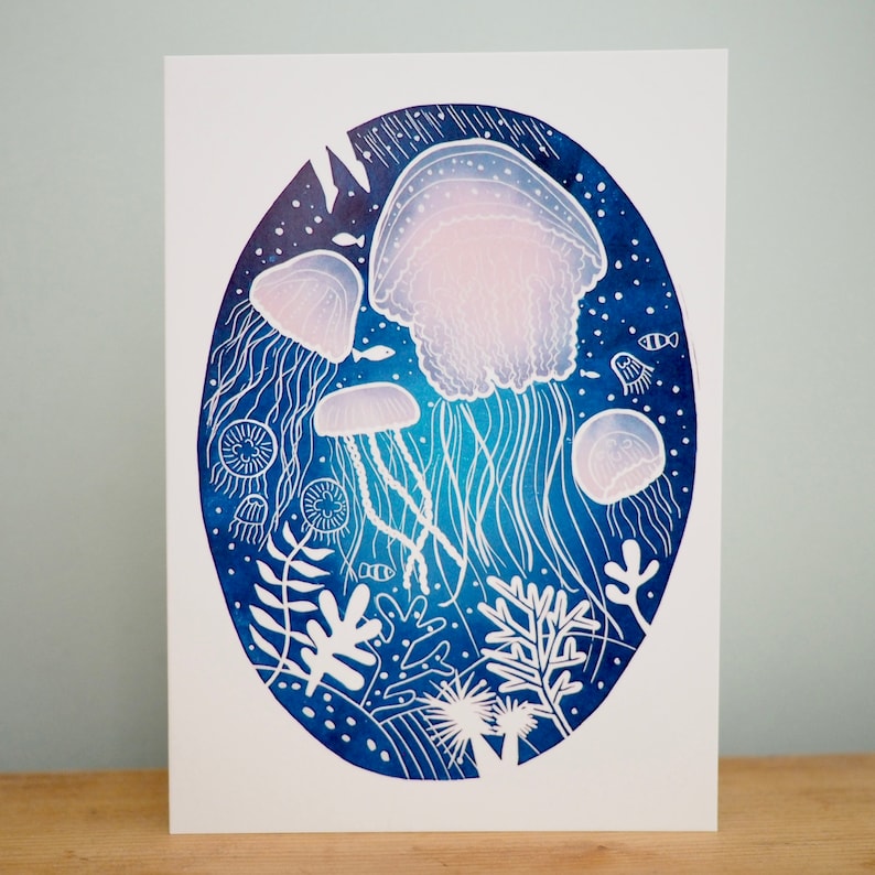 May include: An art print featuring a watercolor illustration of jellyfish in a deep blue ocean scene. The jellyfish are depicted in white and light pink, with long, flowing tentacles. The print includes coral and fish.