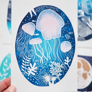 May include: An oval-shaped art print featuring a detailed illustration of jellyfish in a deep blue ocean scene. The jellyfish are white and pink, with long, flowing tentacles. The background includes coral and small fish.