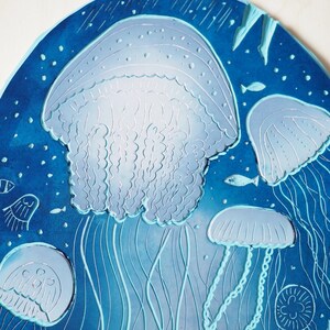 May include: A round, blue decorative piece featuring a detailed illustration of jellyfish. The design includes several jellyfish in varying sizes, with long, flowing tentacles and a textured appearance. Small fish and bubbles are also included.