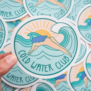 May include: A round sticker with a turquoise blue background and a white border. The sticker features a stylized illustration of a person diving into a wave with a sun and clouds in the background. The text "COLD WATER CLUB" is written in a turquoise blue font around the image.