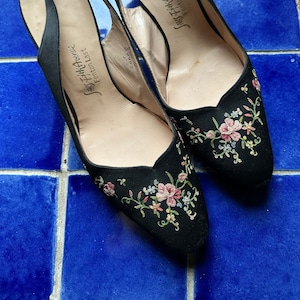 Puede incluir: Un par de tacones de satén negro con tiras en el empeine y bordados florales. Los tacones están decorados con flores rosas, amarillas y verdes. Los zapatos están etiquetados "Sally Thakavore Fenton Last"