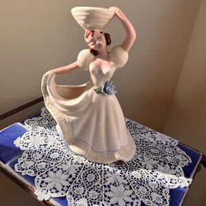 Op de afbeelding: Vintage keramieken beeldje van een vrouw in een vloeiende jurk, die een mand op haar hoofd balanceert. Het beeldje is geschilderd in zachte roze en witte tinten, met een blauw bloemaccent. Het beeldje wordt tentoongesteld op een blauw tegeloppervlak met een wit kanten kleedje.
