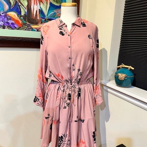 May include: A pink floral print dress with long sleeves, a button-up front, and a cinched waist. The dress has a tiered skirt and a black floral pattern.