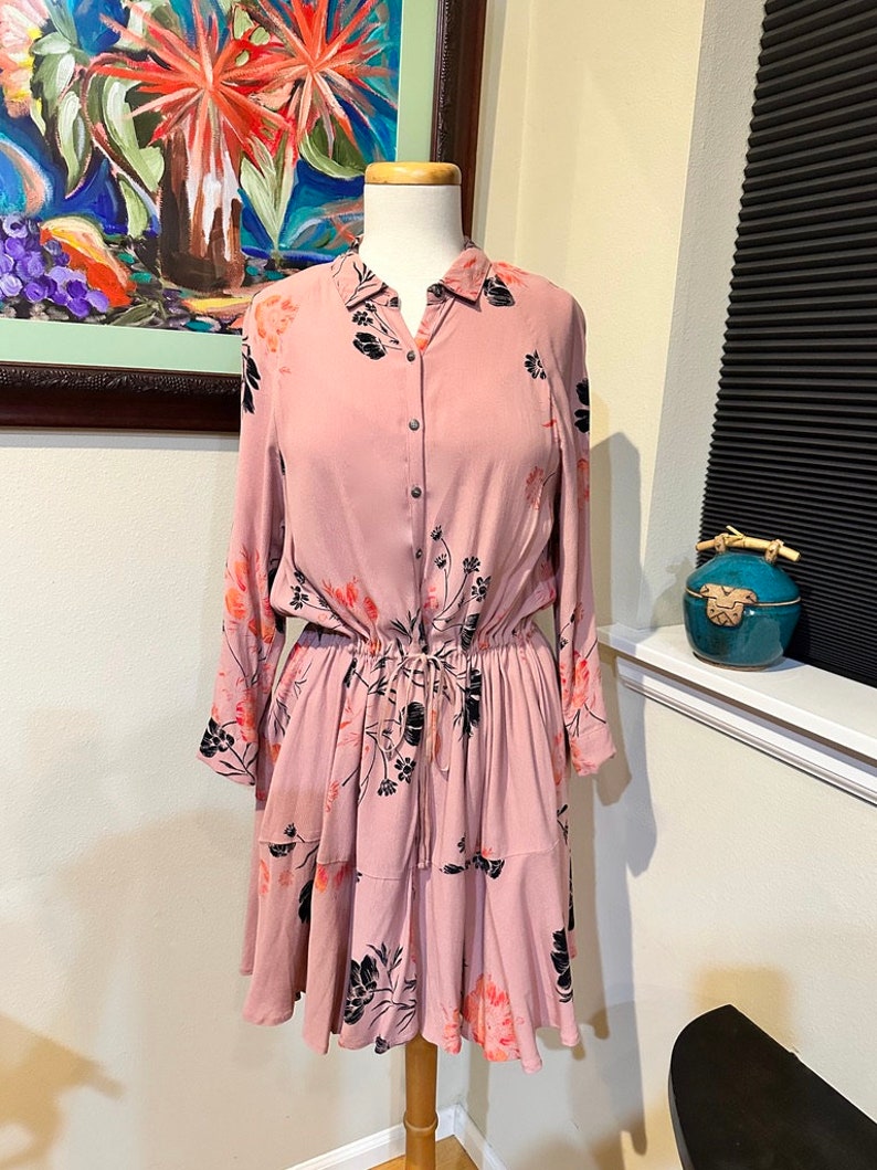 May include: A pink floral print dress with long sleeves, a button-up front, and a cinched waist. The dress has a tiered skirt and a black floral pattern.