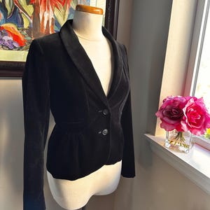 以下が含まれることがあります： マネキンに展示された黒いベルベットのブレザー。 ブレザーは、ノッチ付きラペル、2つボタン留め、フィットしたシルエットのクラシックなデザインです。 ジャケットは絵画と花の咲く窓の前に展示されています。