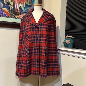 Könnte beinhalten: Ein roter, schwarzer und grüner karierter Umhang mit Knopfverschluss und zwei Taschen. Der Umhang befindet sich auf einem Mannequin und wird vor einer weißen Wand präsentiert.